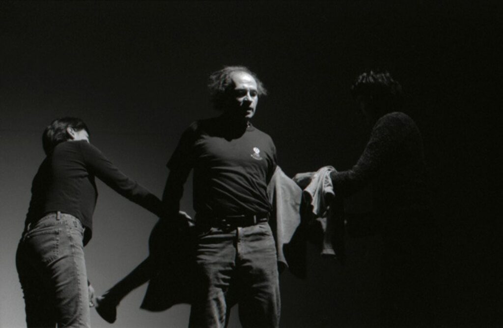 A person under a spotlight has their jacket taken off by two other people, both of whose faces are shrouded in shadow.