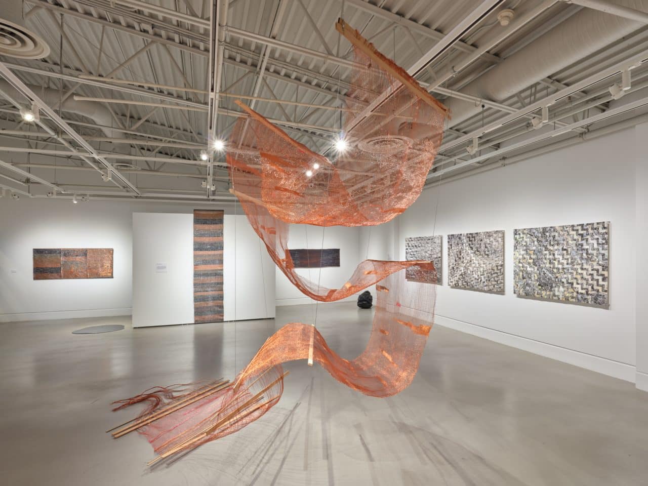 Installation view of a gallery space showing a large suspended copper mesh sculpture that twists through the room, surrounded by woven textile pieces mounted on the walls.
