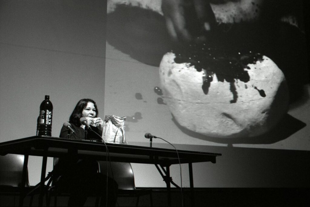 A person eats food at a folding table which bears a bottle of water, three microphones and a McDonald’s paper bag against an image of a hand digging into a white sphere projected onto the wall behind them.