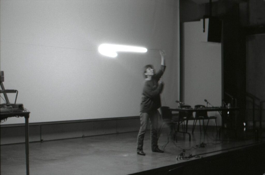 A black and white image depicting a person on a stage swinging a light around on a string above their head.