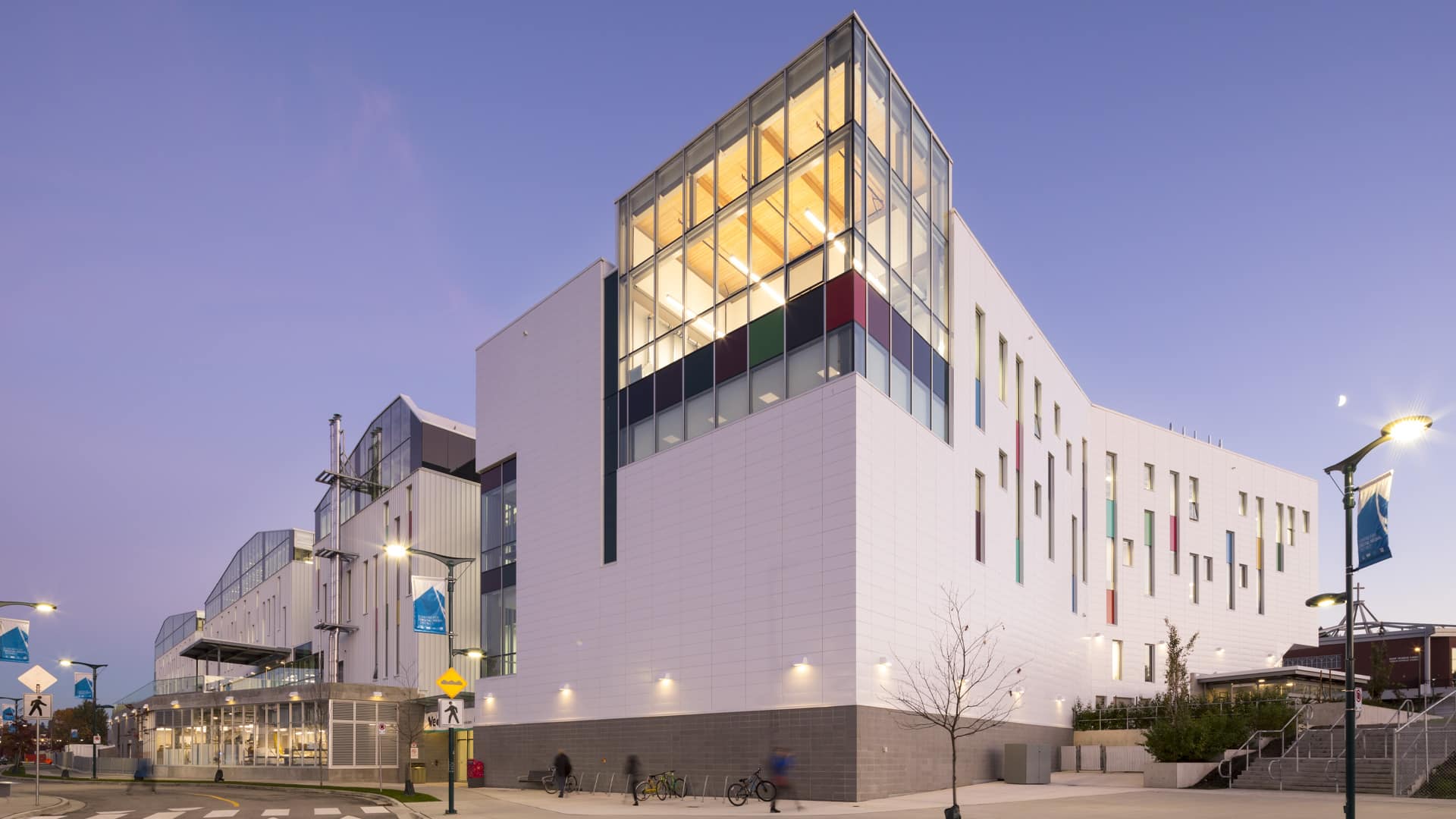 The modern white facade of Emily Carr University of Art + Design glows at dusk under a deep blue twilight sky. Large glass windows reveal warmly lit interior spaces with visible wooden ceilings and colorful panels in red, green, and blue. Students move along the sidewalk below, some walking, others near parked bicycles. Streetlights are on, casting soft pools of light on the pavement, while banners hang from lampposts and a crosswalk sign is visible at the corner.