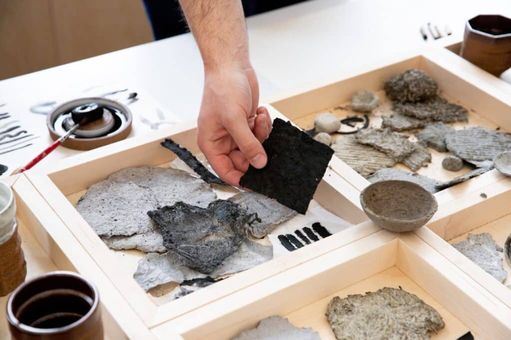 A hand lifting a dark, fibrous sheet from a tray filled with layered handmade material samples, highlighting the rough textures and mixed natural materials.