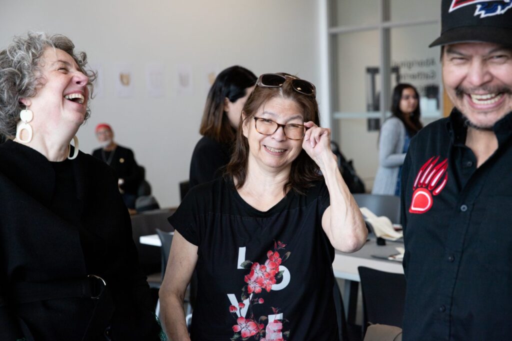 People share a lighthearted moment in a bright studio space. Three individuals stand close together, smiling and laughing. One person adjusts their glasses while another wears white sculptural earrings. A third person in a black shirt with a red bear-paw emblem grins widely. Other participants and artworks are visible softly in the background.