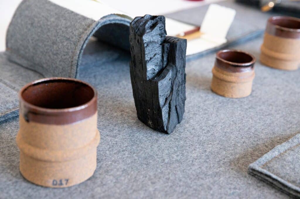 A charred wood fragment standing upright on a felt-covered table beside ceramic vessels and tools used for material exploration.
