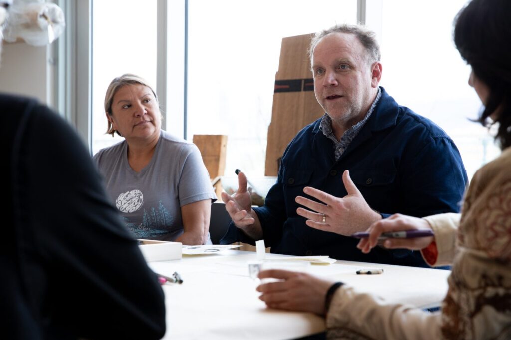 Two workshop participants in conversation at a shared table, surrounded by papers, samples and drawing materials.