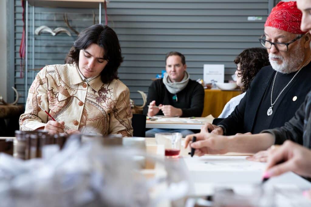 Participants seated at a long studio table painting and drawing together, with brushes, inks and textured samples spread across the workspace.