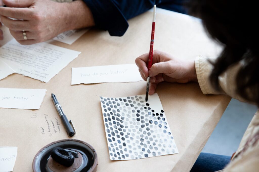 A close view of a hand painting a grid of black ink dots on textured paper, surrounded by handwritten notes and drawing tools.