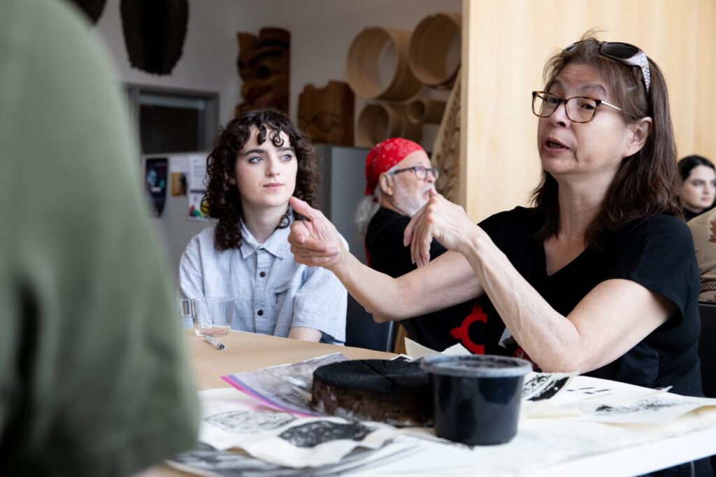 Participants gathered around a workshop table covered with prints, inks and material samples as one person gestures while speaking.