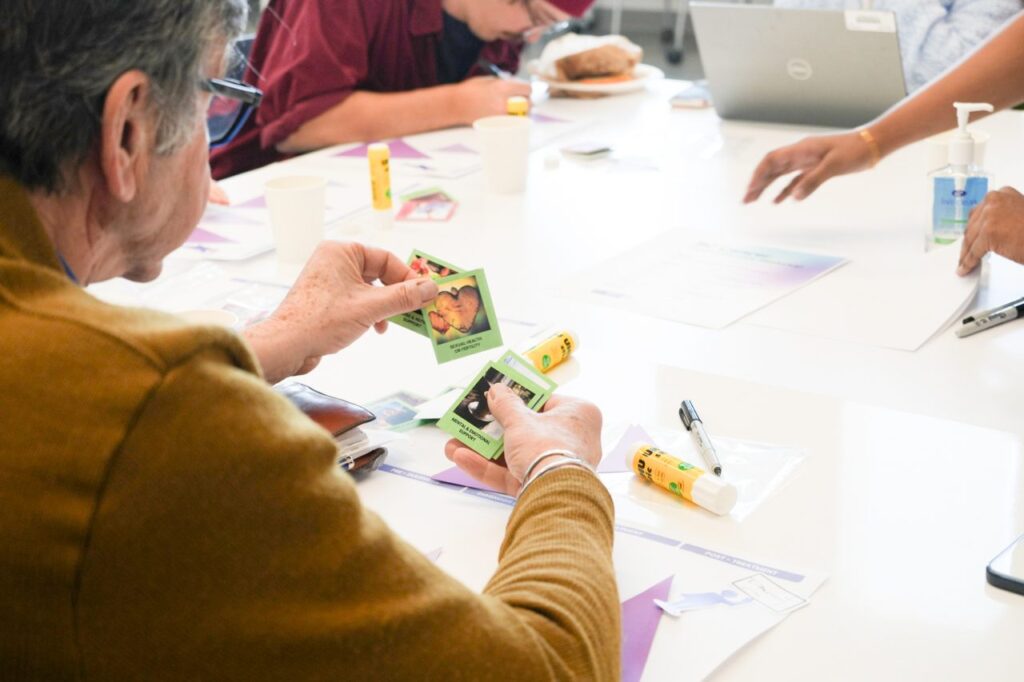 A participant holds several small green cards printed with photos during a table activity. Glue sticks, pens and workshop worksheets are spread across the white tabletop.