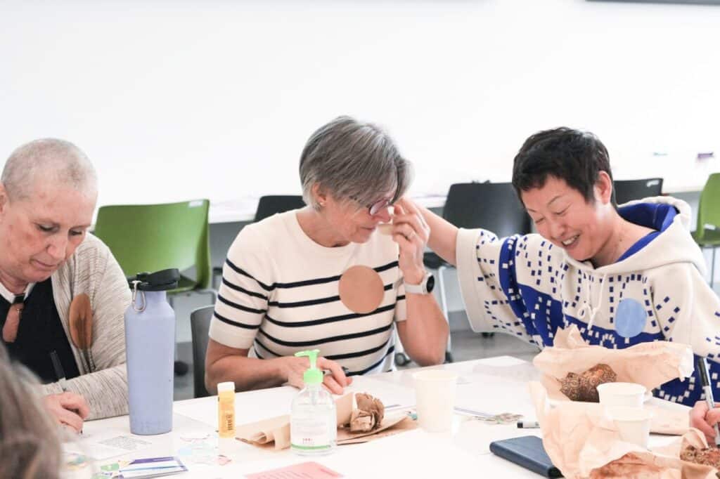 Participants sit together at a workshop table covered with papers, cups and art materials. One person wipes their eyes while another gently reaches over with a supportive touch. A third participant focuses on writing, creating a quiet moment of care and connection in the group setting.