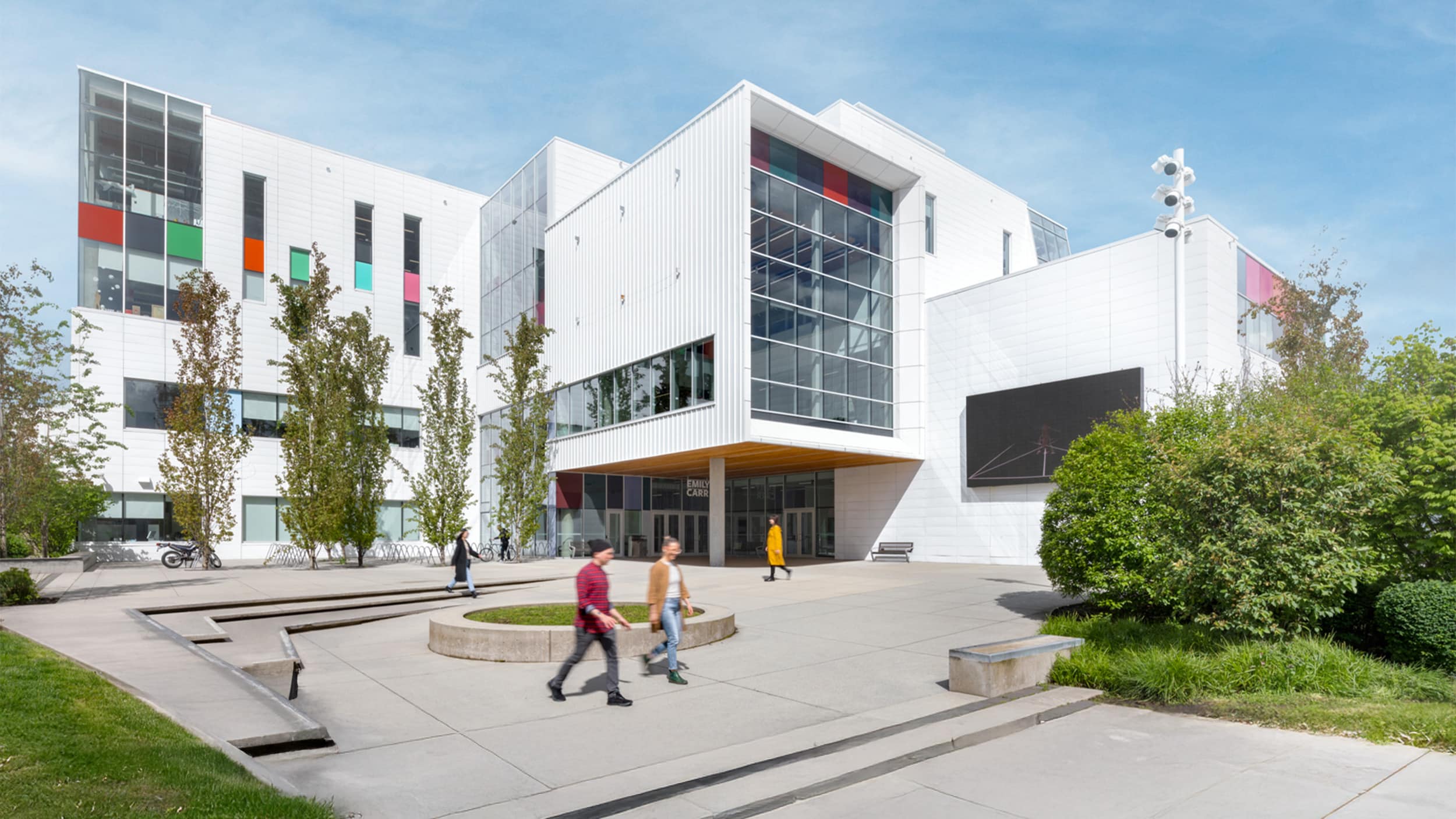A bright, sunny day at the modern campus of Emily Carr University of Art + Design. The white, geometric building features large glass windows, colorful accent panels in red, green, and orange, and a prominent entrance with a yellow overhang. Students walk across the paved plaza, which includes landscaped planters, trees, and concrete benches. A large digital screen is mounted on the right wall, and bicycles are parked near the entrance.