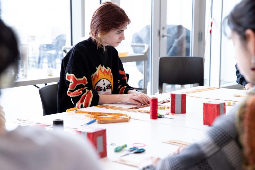 A person in a black sweatshirt works with cedar strips while they watch another person demonstrate weaving techniques.