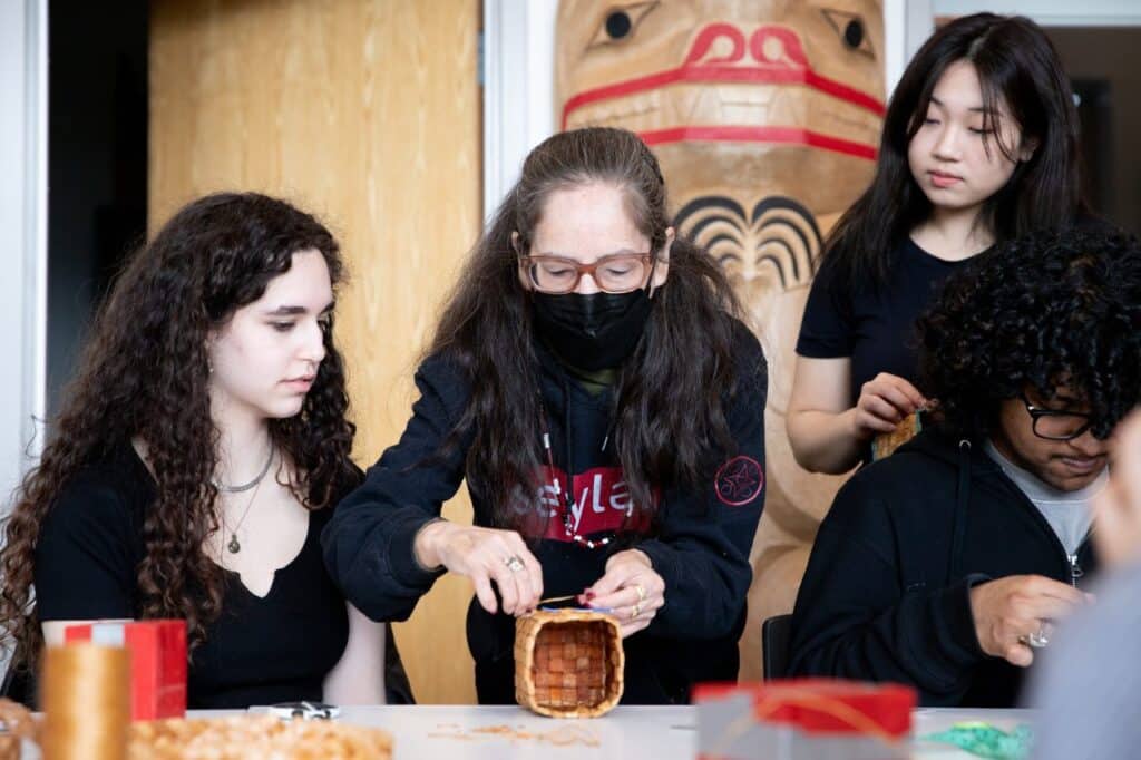 Two people watch a third person attach an abalone button to a woven cedar basket.