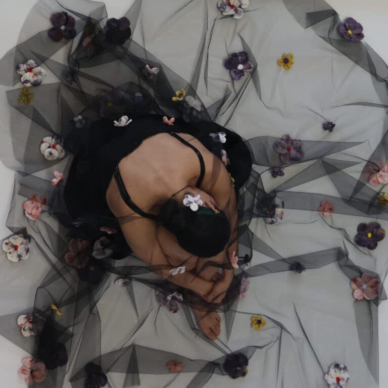 A person sits curled inward on a floor-length black tulle skirt that fans out around them. The sheer fabric is dotted with small, colourful felted flowers, creating a soft halo effect as the figure rests with their head bowed and hands tucked close.
