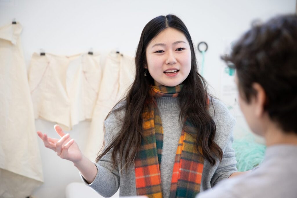 A person with dark hair wearing a plaid scarf and grey sweater speaks with another person and gestures toward a series of linen textiles hanging on the wall.