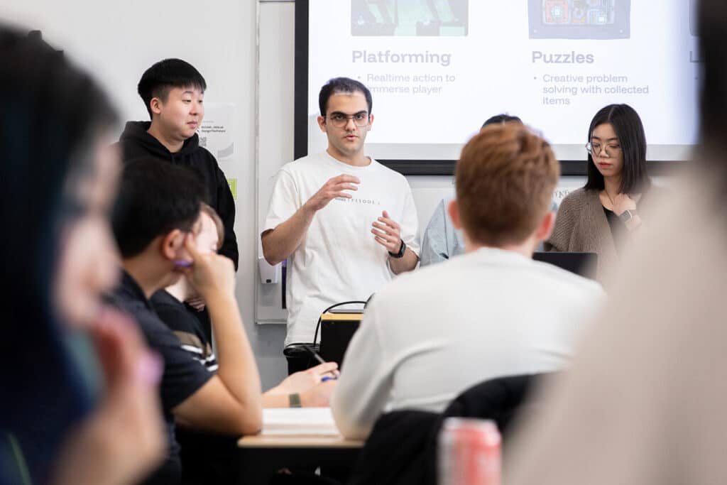 In front of a projection screen, four people stand in a row presenting to a seated audience in a white room.