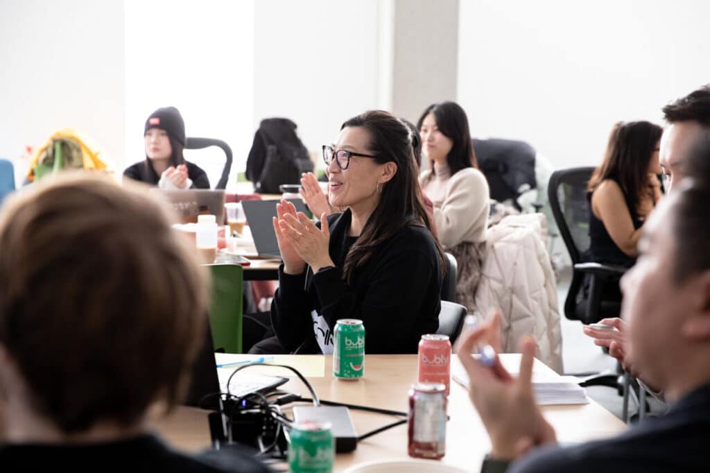 A person seated amongst an audience in a brightly lit room smiles and applauds.