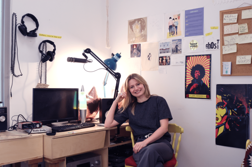A person in a striped shirt sits in a wooden chair leaning on a desk covered in recording equipment in a room whose walls bear posters of famous musicians.