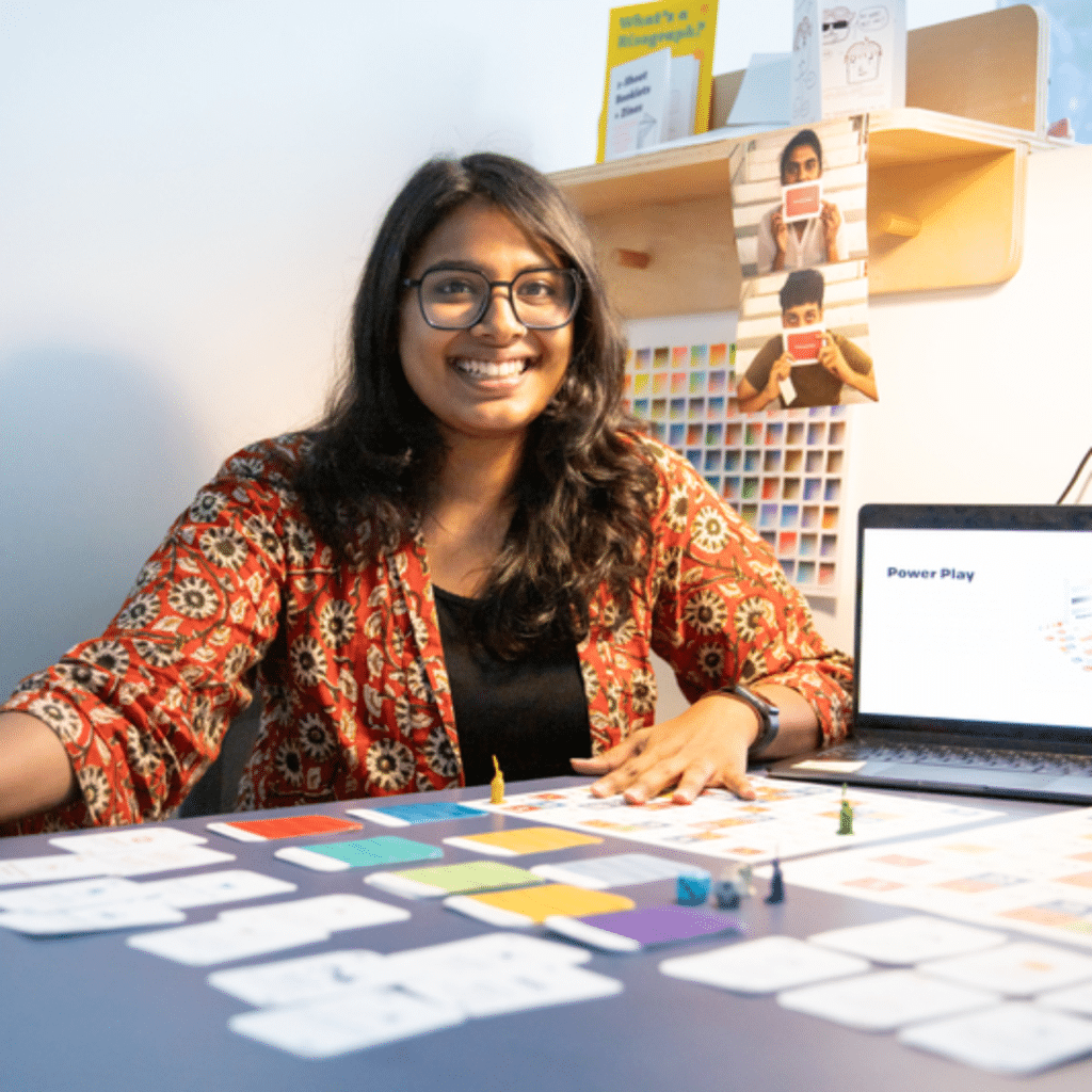 A person sits at a table covered in colourful game cards, tiles and small figurines, smiling toward the camera as they demonstrate their design project. A laptop displays a slide titled “Power Play” and printed colour swatches and photos hang on the wall behind them.