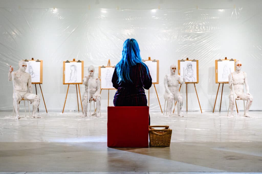 In a white room with plastic on the floor and walls, a person with blue hair and wearing a purple robe sits on a red block with a wicker basket on the floor to the right and their back turned while facing four plaster-covered individuals, each with white staves in one hand, who are seated on stools next to drawings on easels of the person with the blue hair.