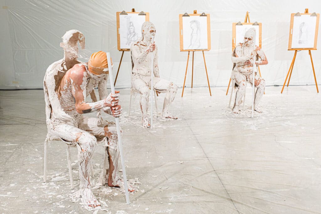 In a white room with plastic on the walls and floor, a person holding a white staff to the right of frame leans forward to break free from a shell of thin plaster in which they were encased, while two others in the centre and left of frame struggle to do the same. Visible in the background are life drawings on easels.