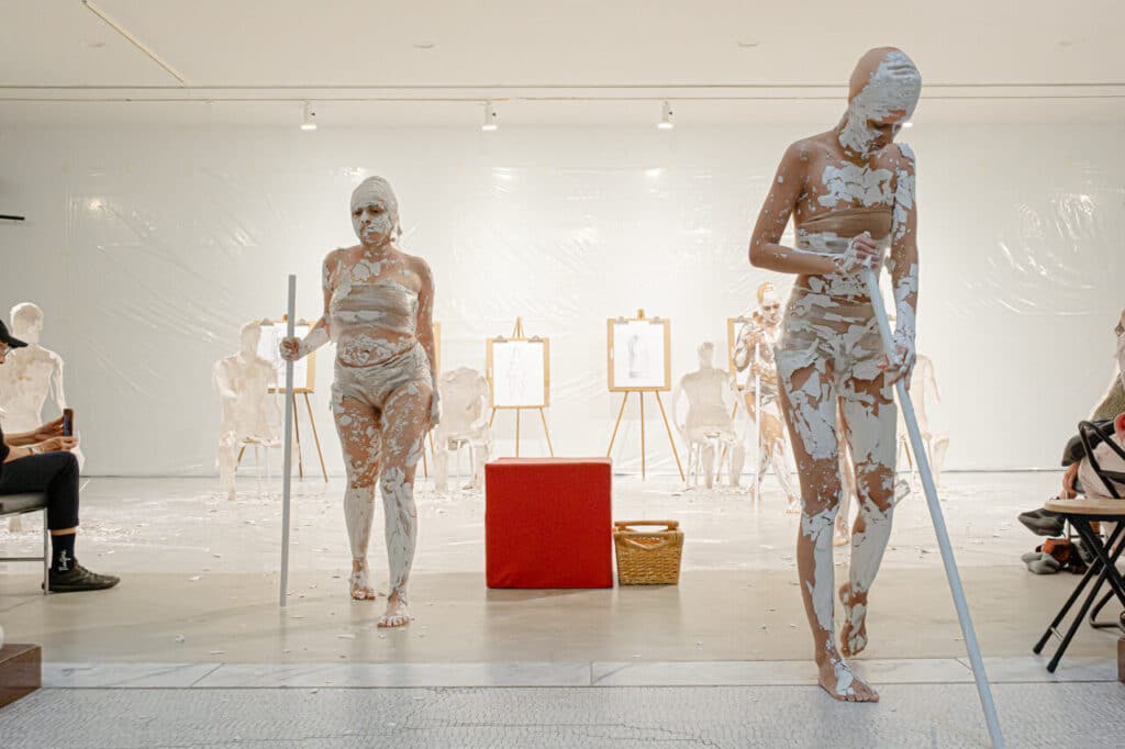Two people leaning on staves and whose bodies are covered in cracked, dried plaster walk out of a plastic-draped room while in the background can be seen plaster shells from which they emerged and in between them is a red block and a wicker basket.