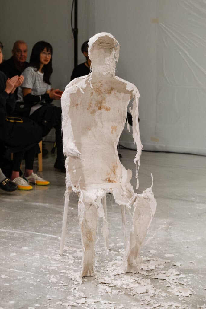 People in the background applaud while in the foreground rests a plaster shell in the shape of a person seated on a chair.