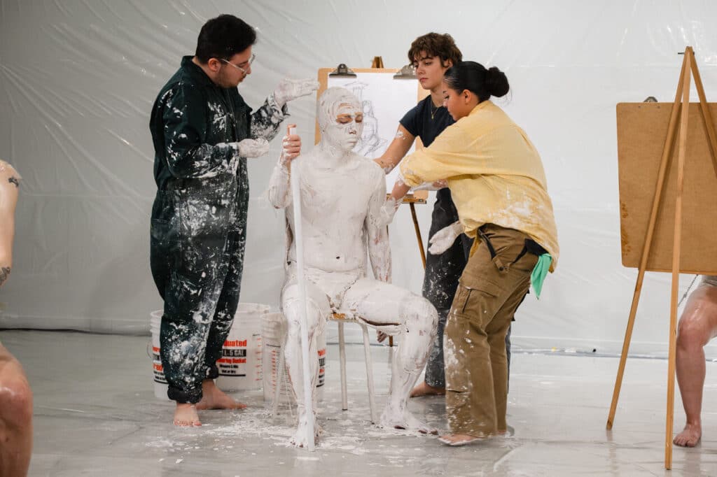 In a white room with plastic on the walls and floor, a seated person wearing an orange swim cap and with a white staff in their hand is smeared with plaster by three other people while in the background and off to the sides people can be seen executing life-drawings.