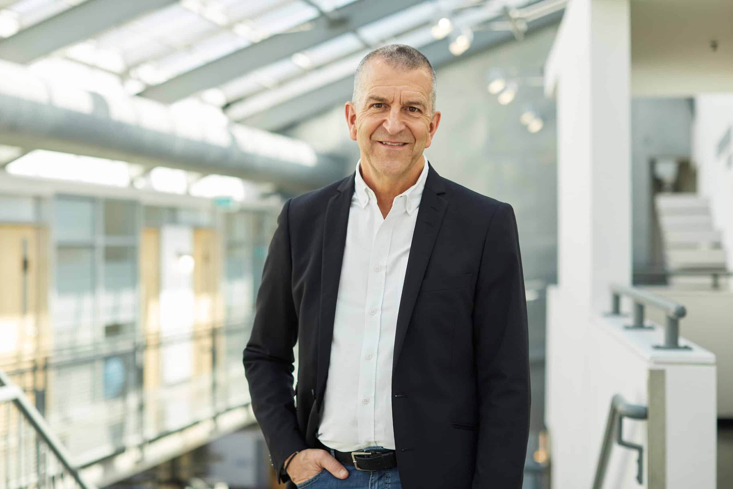 A person in a white shirt and dark blazer smiles in a room with sun pouring through skylights.