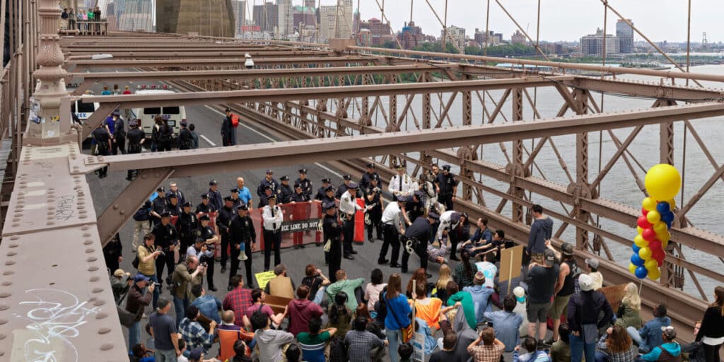 Crowd gathered on a bridge roadway as police block traffic, with city buildings visible in the background.