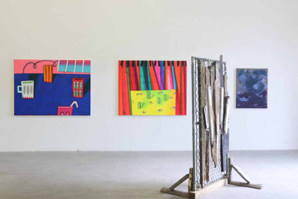 A series of colourful paintings adorn a white wall while in the foreground stands a sculpture made from ragged pieces of found wood affixed to a section of chain link fencing and mounted to old two-by-fours.