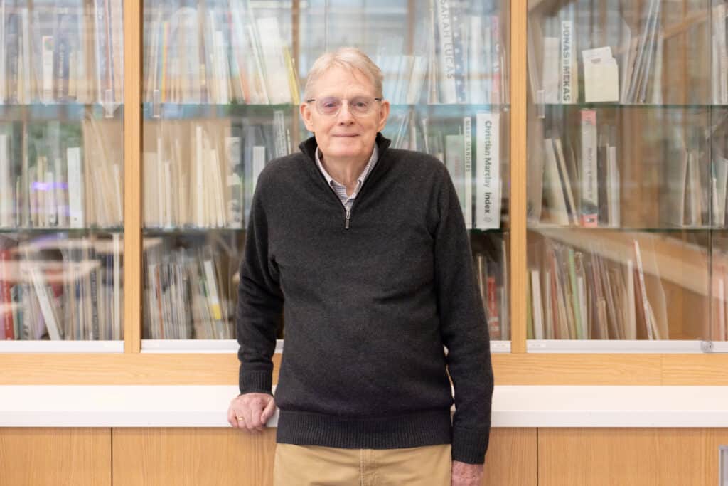 A person wearing glasses and a dark sweater stands indoors in front of a glass-fronted bookshelf filled with books, resting one hand on a counter.