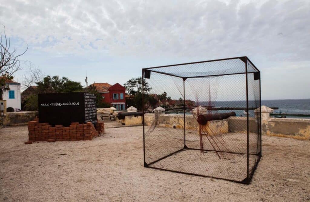 Outdoor installation with a wire cage holding a cannon near the ocean, with brick and stone structures nearby.