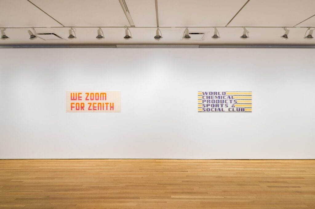 A pair of textile artworks hang on the wall of a gallery, the left with text saying "We Zoom for Zenith," the right with text reading "World Chemical Products Sports & Social Club." 
