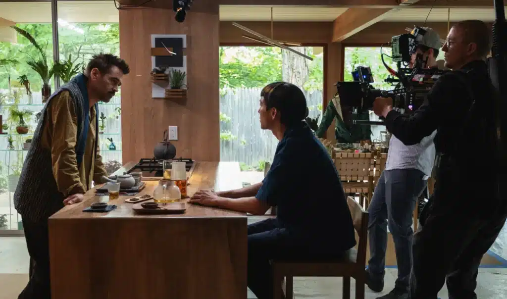A film crew records a scene in a modern kitchen as two people face each other across a wooden table set with tea, while a camera operator films from the side.