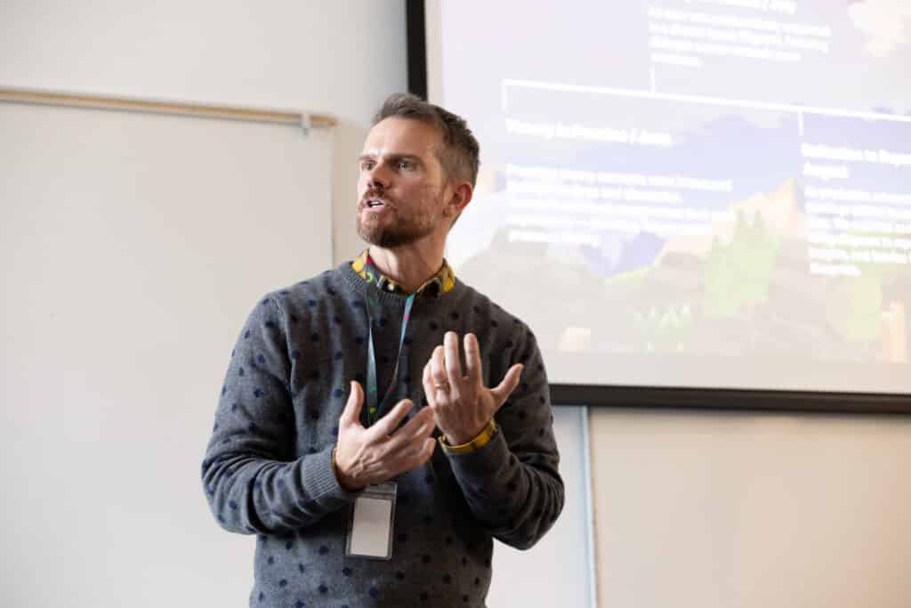 A person in a slate blue sweater talks animatedly in a room with a projection screen on the wall.