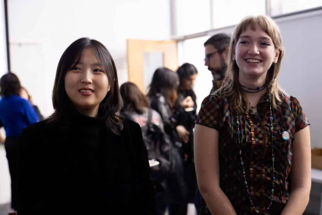Two people in dark clothing smiling in a crowded room, appearing to listen while someone off-camera speaks.