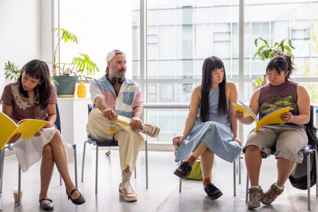 Four people sitting in chairs and some to the right are looking at a yellow book