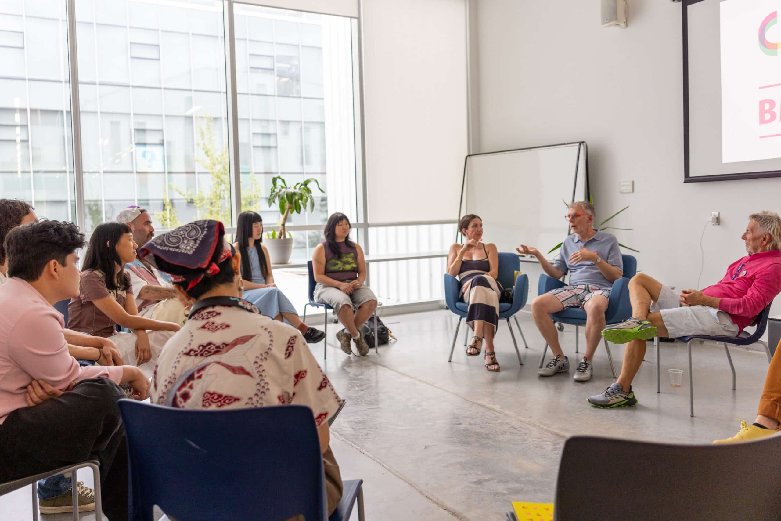 A group of people sit in a loose circle of chairs in a bright studio space, listening and talking during a facilitated group discussion.