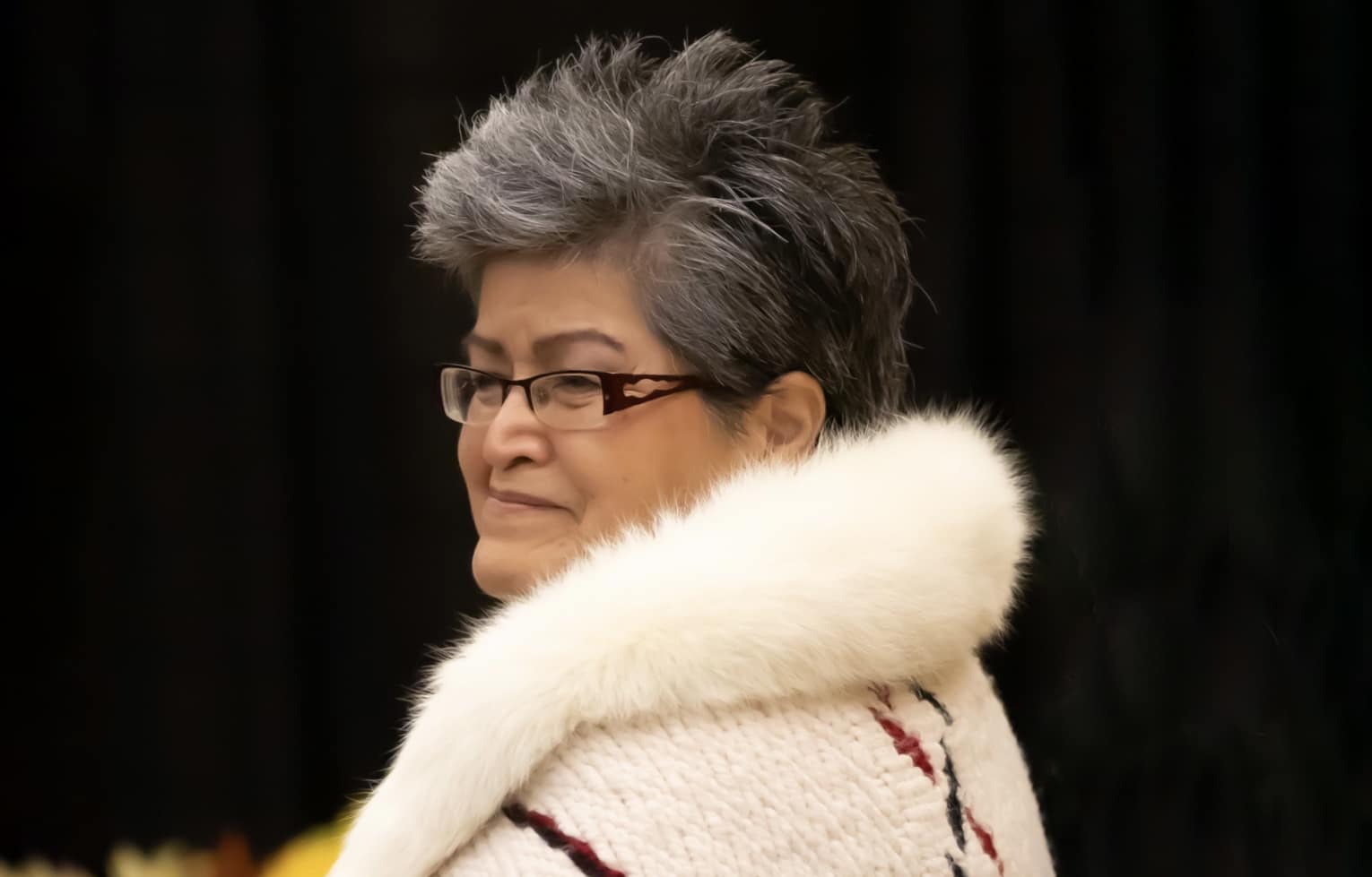 A person with grey hair and glasses wearing a fur-lined, woven coat is visible from the shoulders up against a black background.