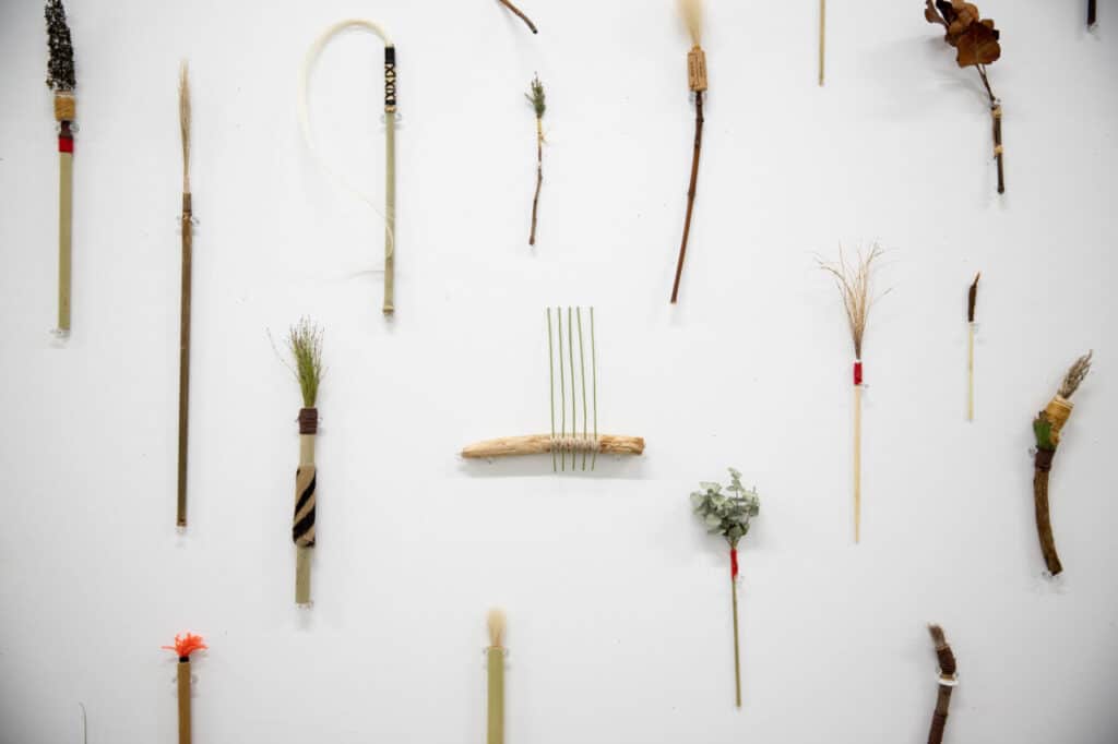 An arrangement of handmade brushes made from natural materials is mounted across a white wall, with each piece spaced evenly to form a grid-like display.