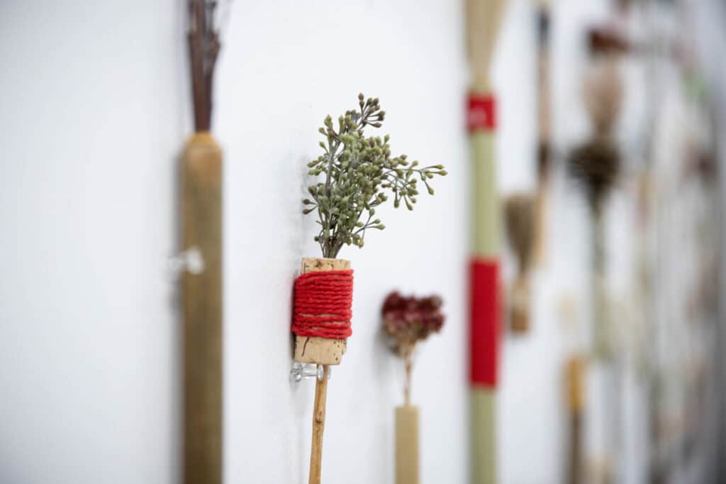 Close-up of a handmade brush mounted on a white wall, constructed from a wooden handle, wrapped red thread, and dried plant material.