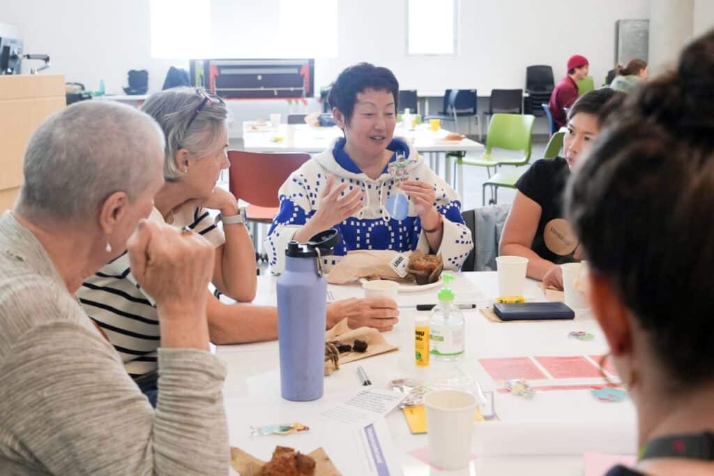 A person in a blue and white hooded shirt sits at a table amongst a group to whom they speak.