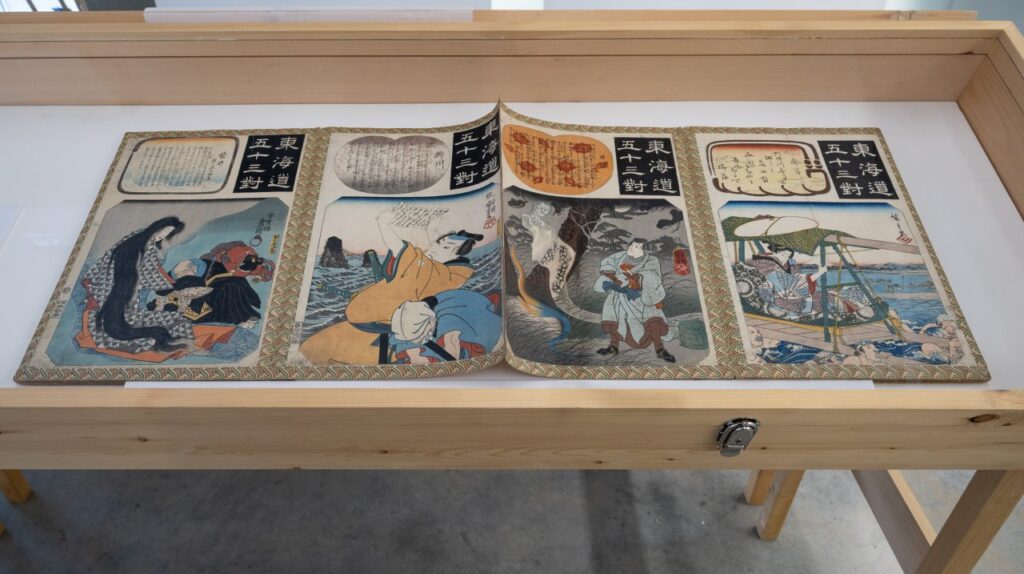 Colourful Japanese prints are displayed in a wooden vitrine with a glass face.