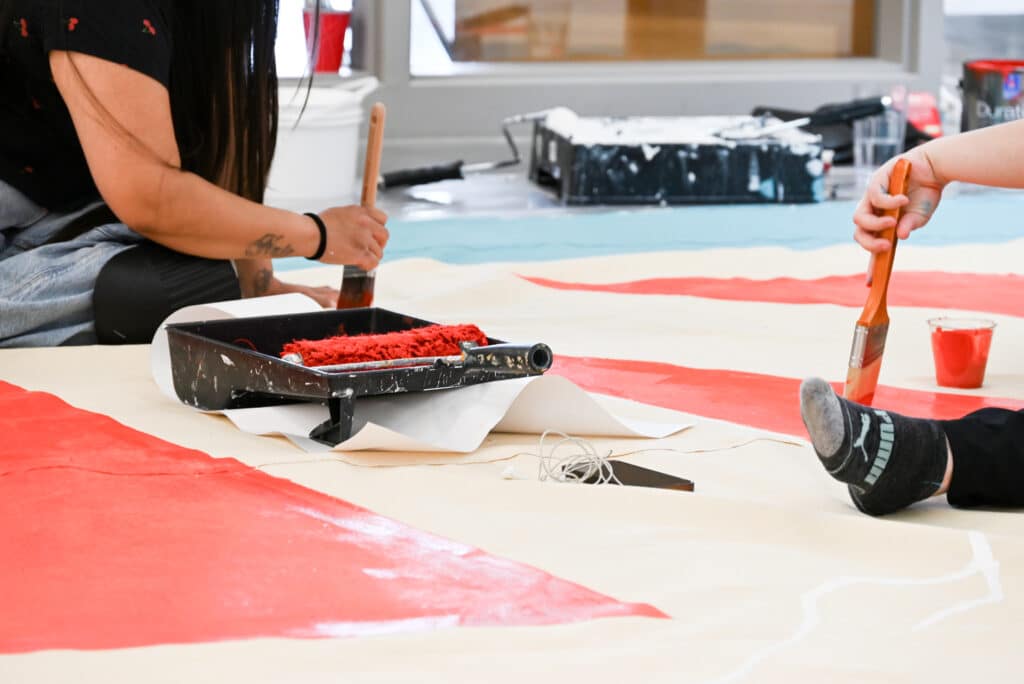 Hands belonging to two separate people hold paintbrushes from which they paint red designs onto a canvas which lays on the floor.