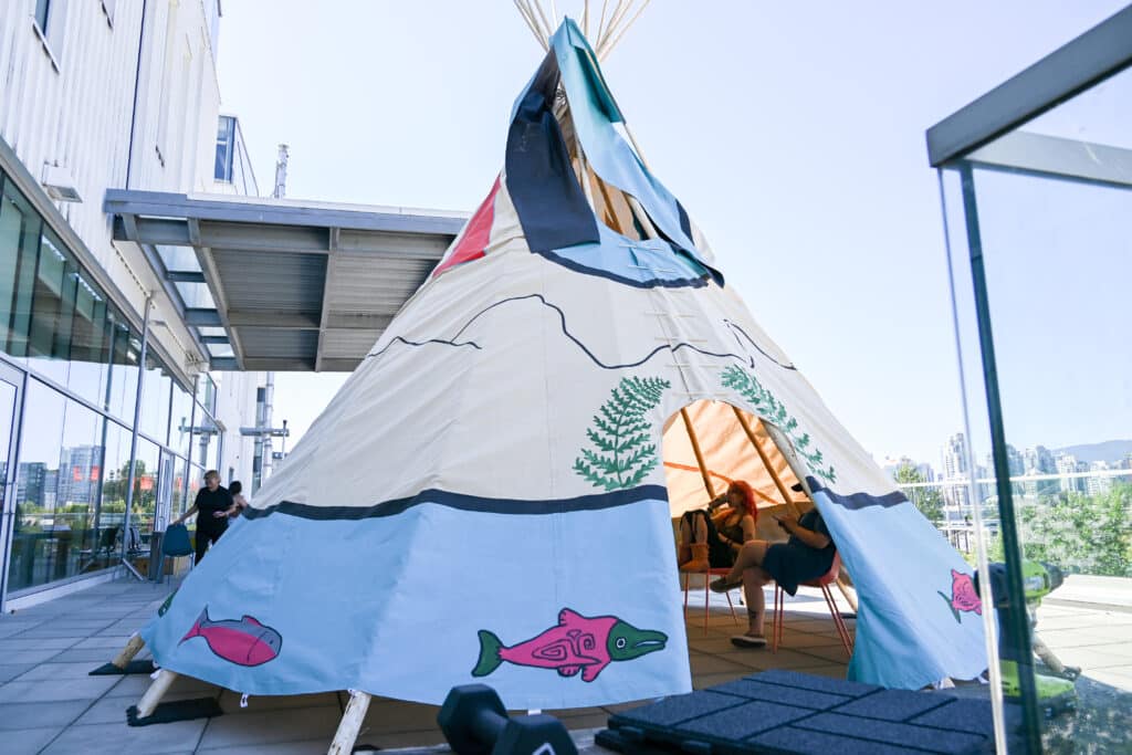 A tipi painted with images of cedar boughs, mountains, sunrays and salmon stands on a concrete patio on a blue sunny day.