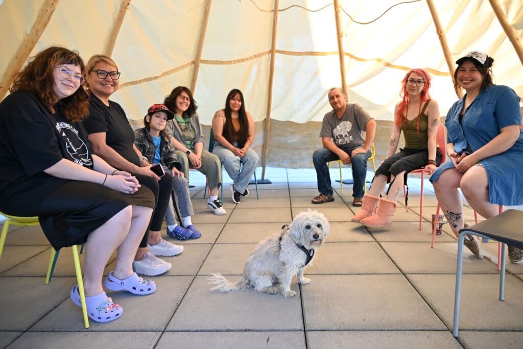 A small white dog is surrounded by a group of people sitting on chairs inside a tipi through whose walls sunlight is filtered yellow.