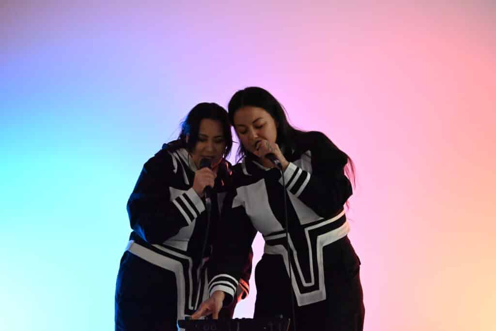 Two people in black clothing with white geometric designs lean close together holding microphones against a blue-and-red-lit backdrop.