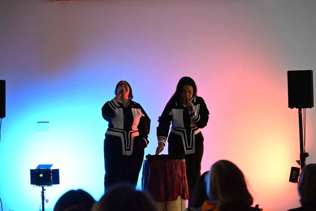 Two people in black clothing with white geometric designs and holding microphones sing for an audience against a blue-and-red-lit backdrop.