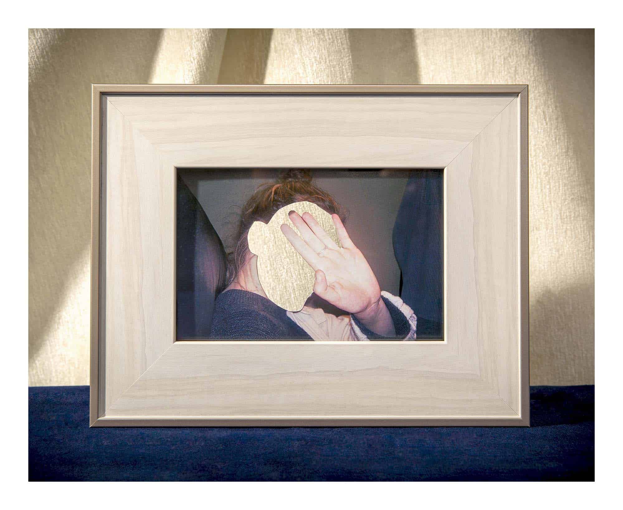 Framed photograph of a person seated indoors with their face obscured by a raised hand and a textured overlay, mounted against a softly lit wall.
